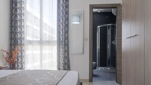 Een hotelkamer met een bed, een badkamer met een douche met glazen wanden en een deur naar een andere kamer.