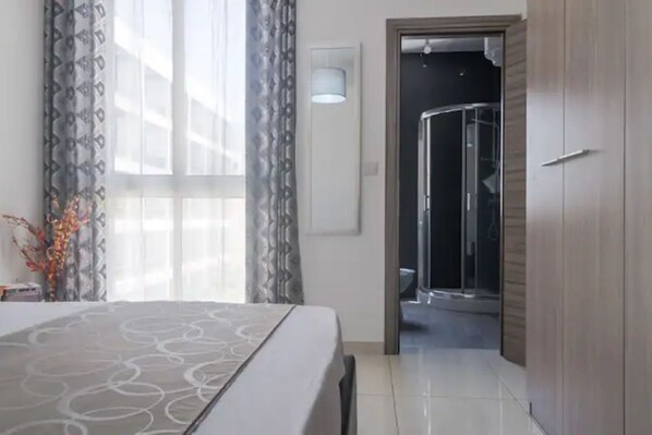 A hotel room with a bed, a bathroom with a glass shower, and a door leading to another room.