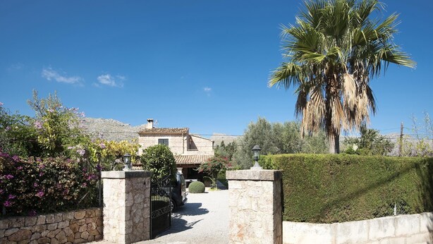 A stone gate leading to a villa with a tiled roof, surrounded by lush greenery and a palm tree. - Finca Can Tomeu, private pool, wifi, 4 bedrooms, 10 minutes to the beach  (Pollenca)
