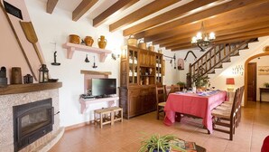 A cozy living room with a fireplace, wooden beams, a dining area with a red tablecloth, and a staircase leading upstairs. - Finca Can Tomeu, private pool, wifi, 4 bedrooms, 10 minutes to the beach  (Pollenca)
