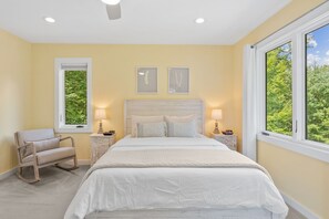 Une chambre à coucher comprenant un lit, deux tables de chevet, un fauteuil à bascule et deux fenêtres donnant sur un paysage verdoyant.