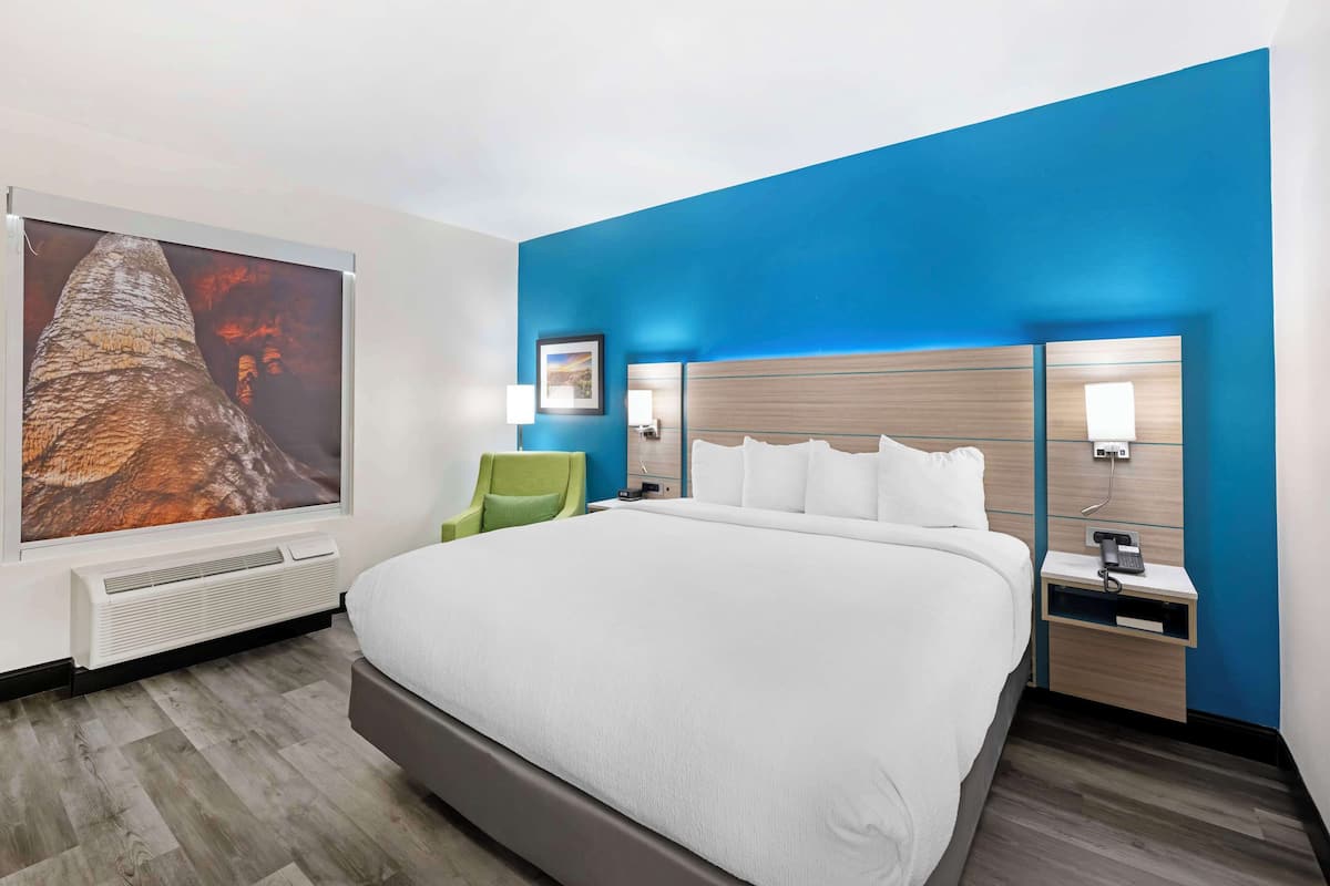 Ein modernes Hotelzimmer mit einem großen Bett, einer blauen Akzentwand, einem Wandbild, einem grünen Sessel und einem Nachttisch mit einem Telefon.