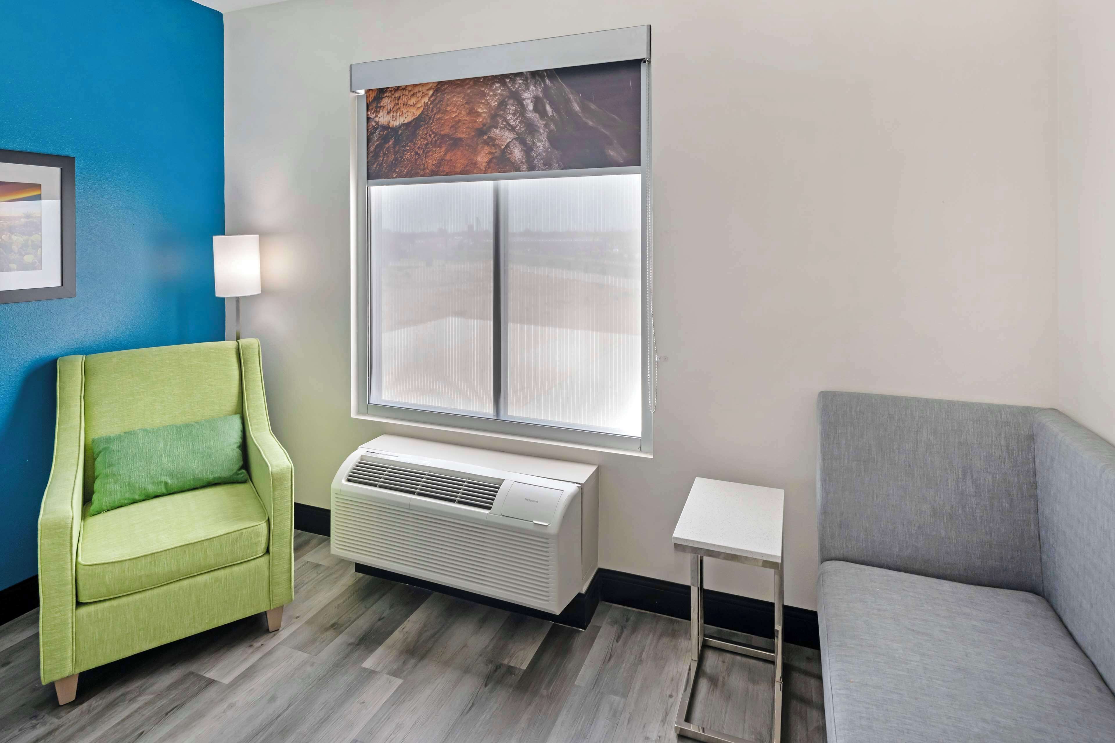 A room with a green armchair, a grey sofa, a small white table, a window with a view, and a wall-mounted air conditioner.