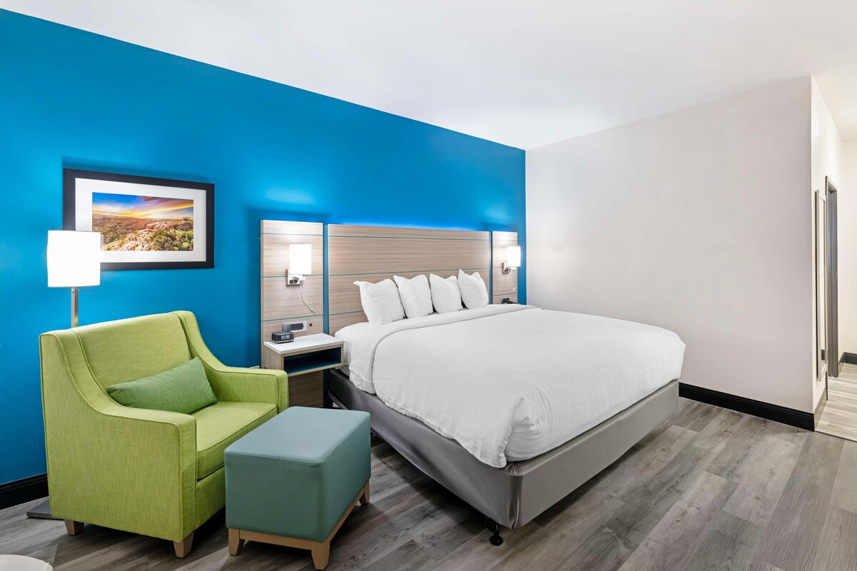 Une chambre d’hôtel avec un lit, un fauteuil vert, un petit pouf, une table de chevet, un tableau encadré au mur et un miroir au-dessus du lit.