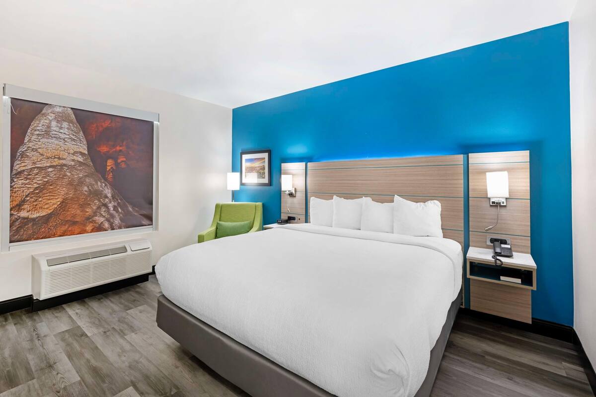 Ein Hotelzimmer mit einem großen Bett, einem grünen Sessel, einem wandmontierten Fernseher und einem Nachttisch mit Telefon.