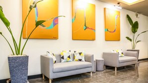 Un hall d’entrée avec un ensemble de canapés gris, de grandes plantes en pot et des tableaux abstraits jaunes accrochés au mur.