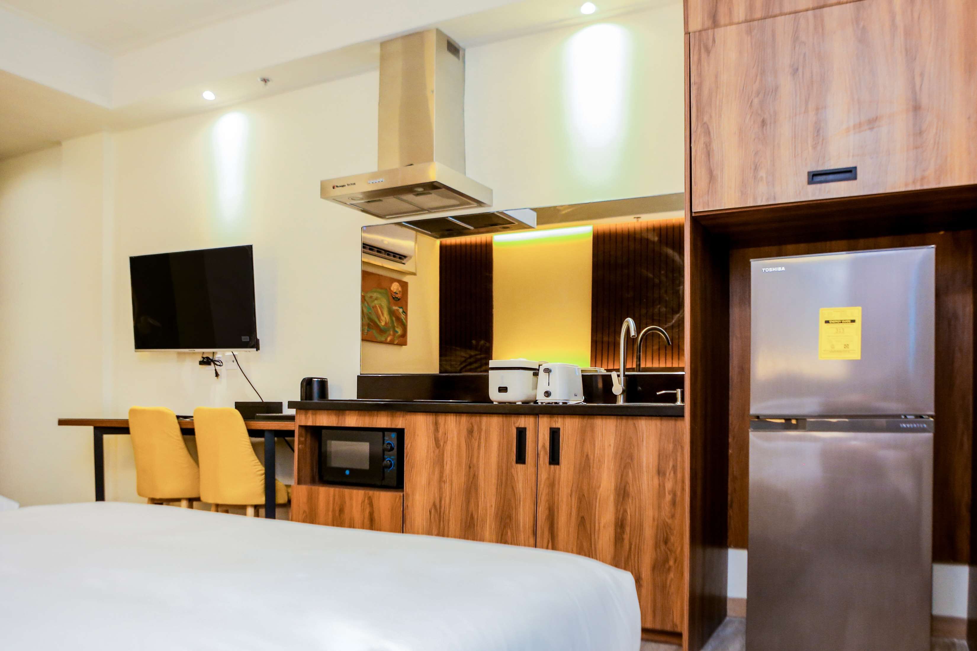 A hotel room with a bed, a desk with a flat-screen TV, a wooden kitchenette with a microwave, toaster, and refrigerator, and a bathroom with a toilet and sink.