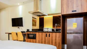 A hotel room with a bed, a desk with a flat-screen TV, a wooden kitchenette with a microwave, toaster, and refrigerator, and a bathroom with a toilet and sink.