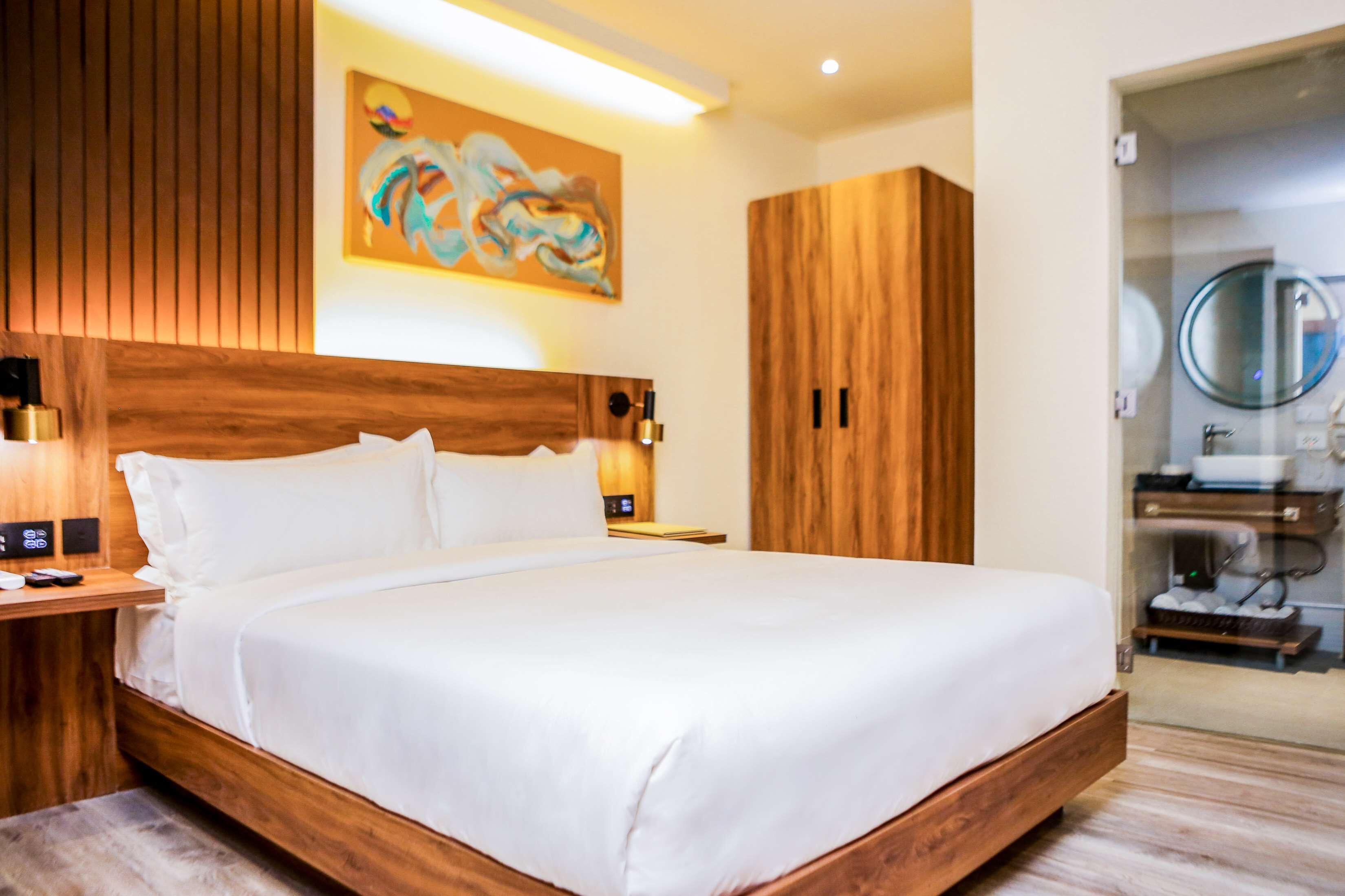 A modern hotel room with a wooden headboard, a large bed, a painting on the wall, and a bathroom visible through a glass door.