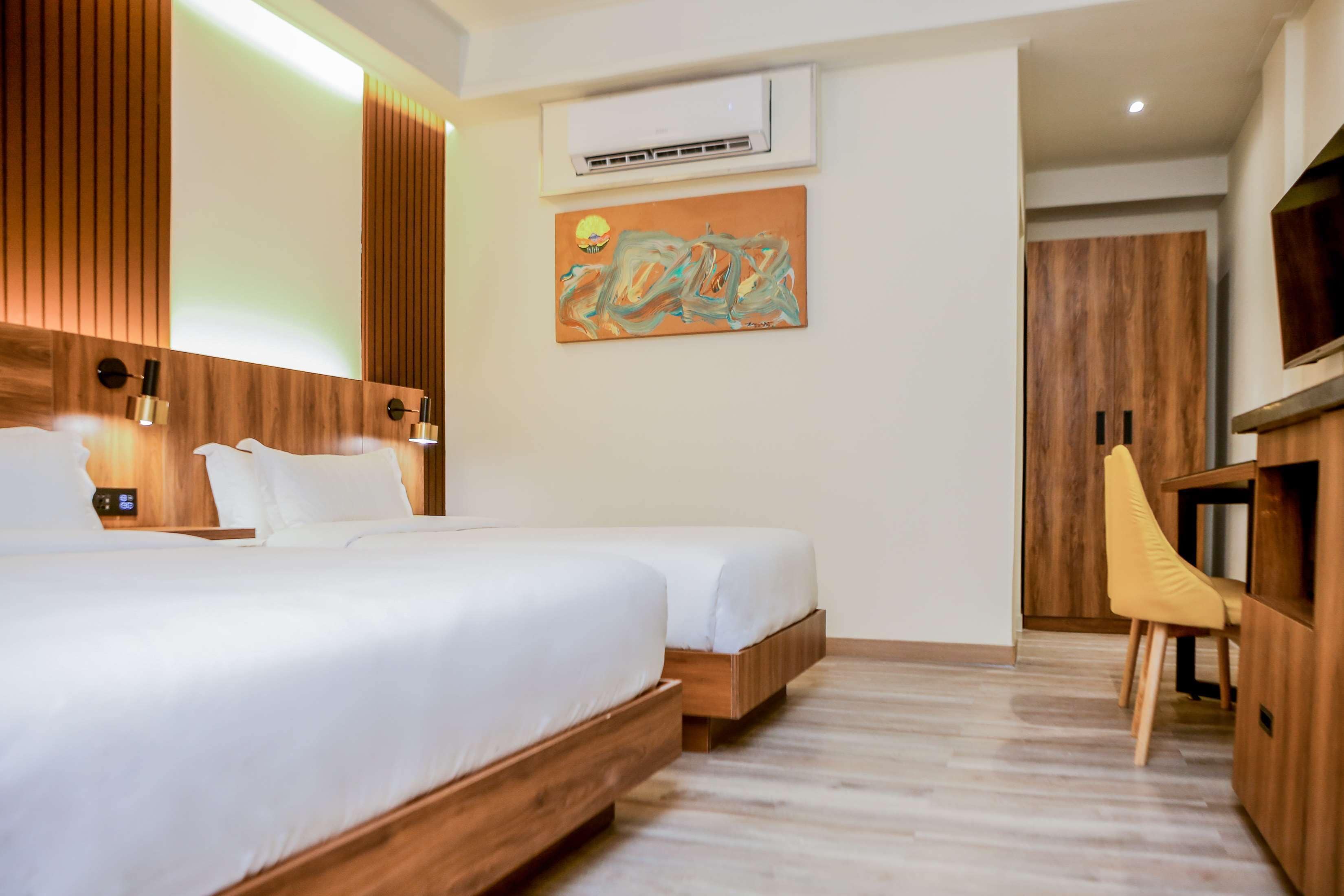 A hotel room with two beds, a wooden headboard, a wall-mounted air conditioner, and a painting.