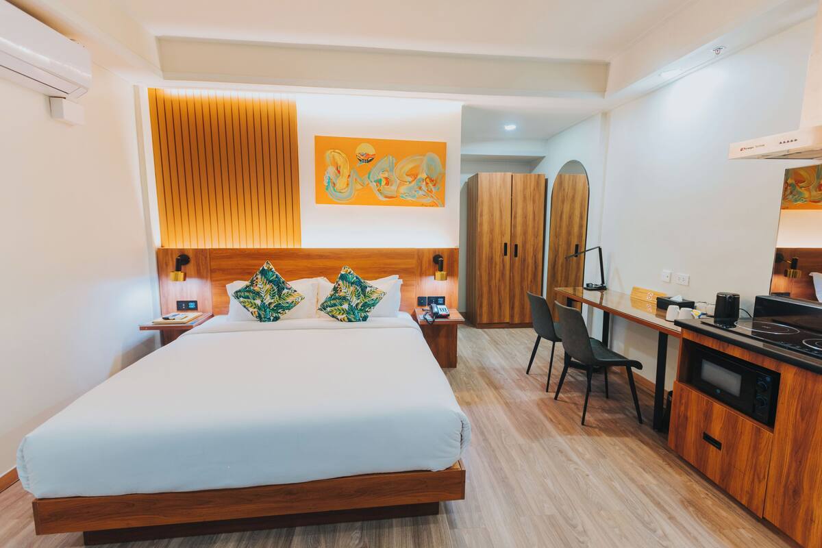 Ein modernes Hotelzimmer mit einem großen Bett, einem Holzkopfende, einem Schreibtisch mit Stuhl, einer Küchenzeile und einem Kleiderschrank.