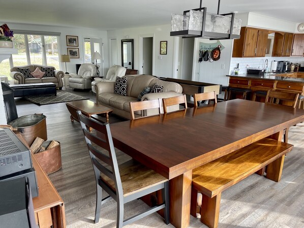 A spacious living room with a large wooden dining table, leather sofas, and a kitchen area with wooden cabinets and a hanging light fixture. - YR ROUND 10 PERSON OUTDOOR HOT TUB! SHUFFLE PUCK, PET FRIENDLY, DESIGNED FOR FUN (Cambridge)