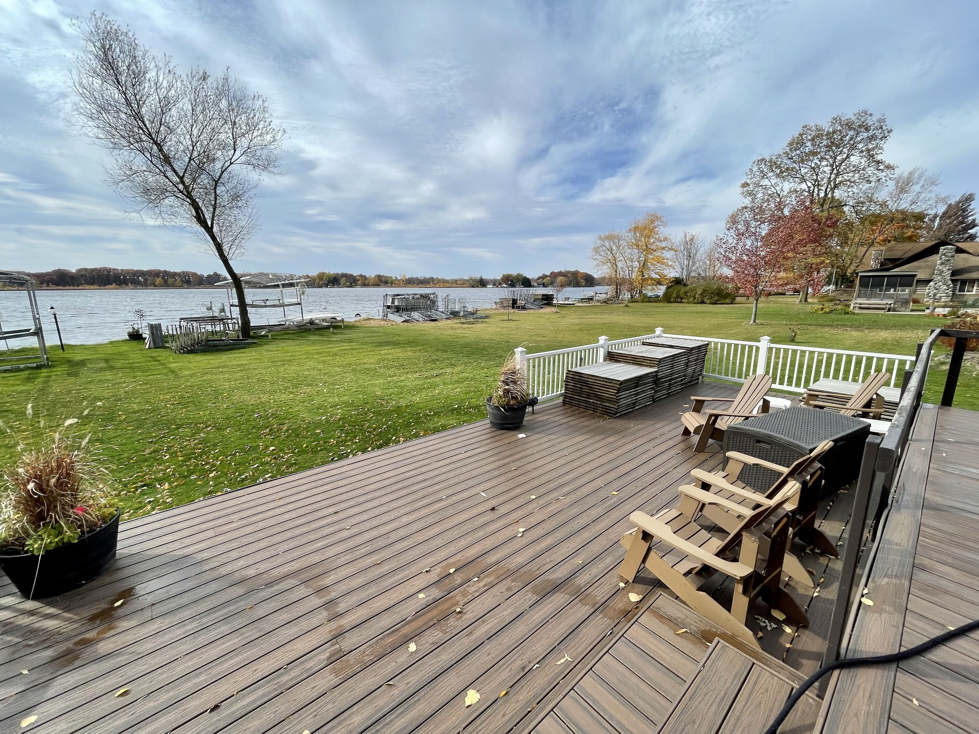 Una terraza de madera con mobiliario exterior, barandilla blanca y vista a un lago.