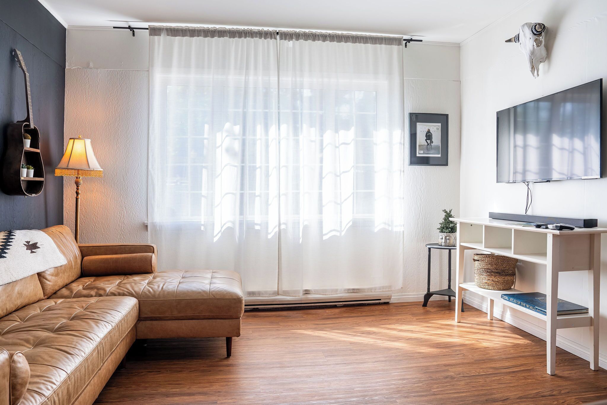A living room with a leather sofa, a standing lamp, a TV on a white shelf, and a guitar on the wall.