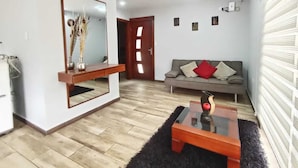 A modern living room with a wooden floor, a sofa with cushions, a coffee table, a mirror, and a door leading to another room. - Welcome to your home in Baños (Baños de Agua Santa)