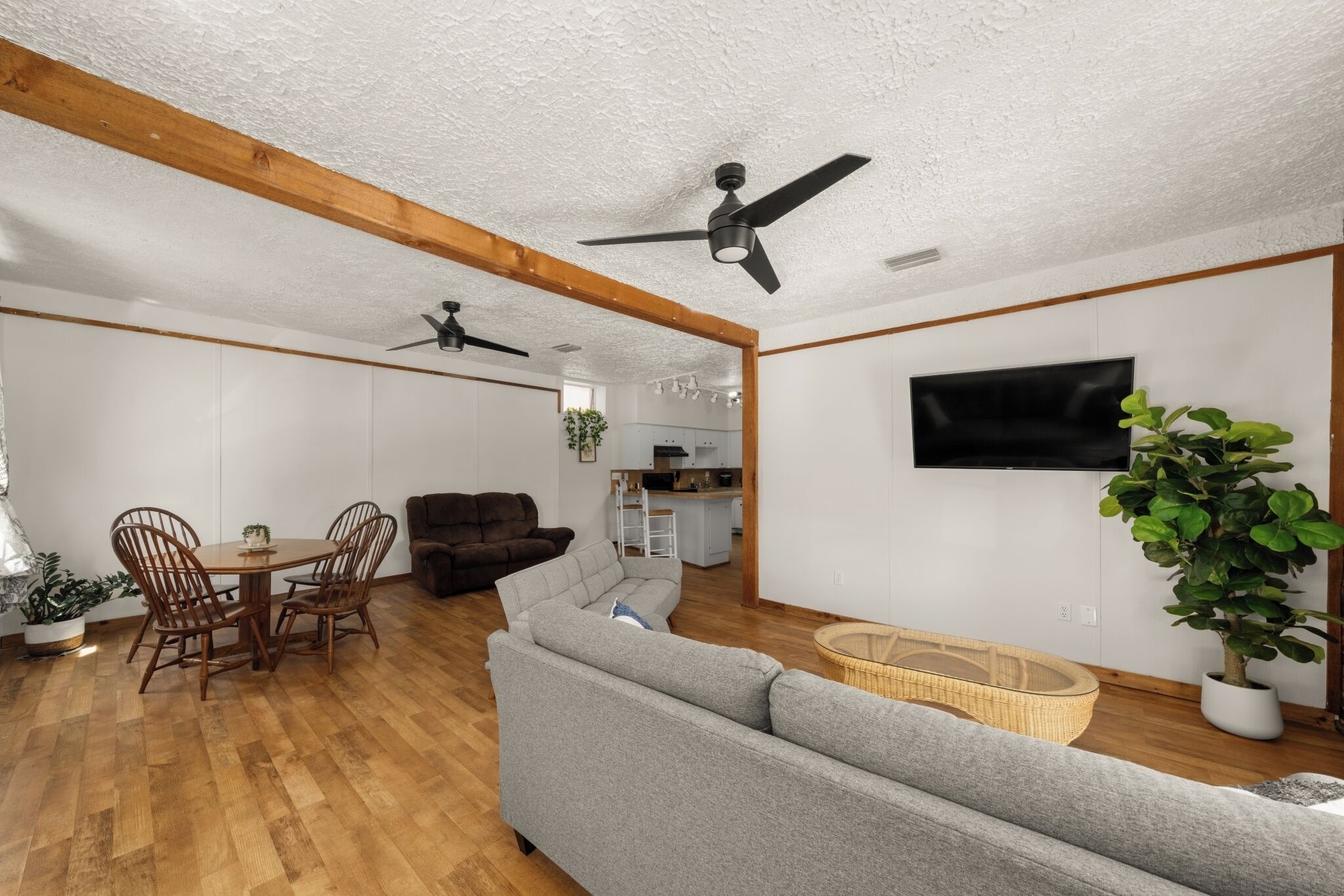A spacious living room with a grey sofa, wooden dining table, and chairs. There is a television mounted on the wall and a ceiling fan.