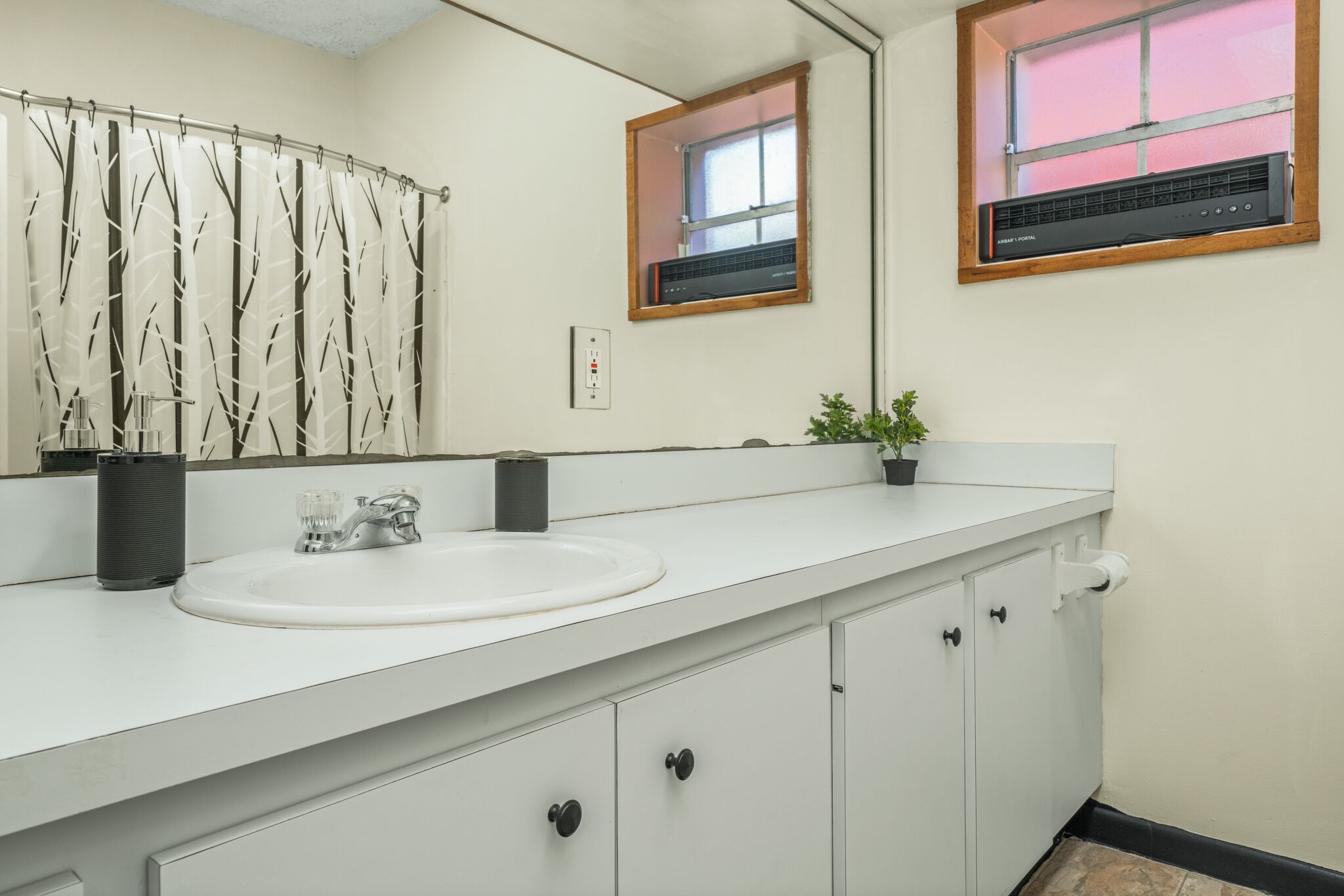 A bathroom with a white sink, a mirror, a shower curtain with a tree pattern, a small window, and a heater on the wall.