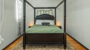 A four-poster bed with a dark wooden frame, green bedding, and decorative pillows. There are two bedside tables with lamps, and a ceiling fan above.
