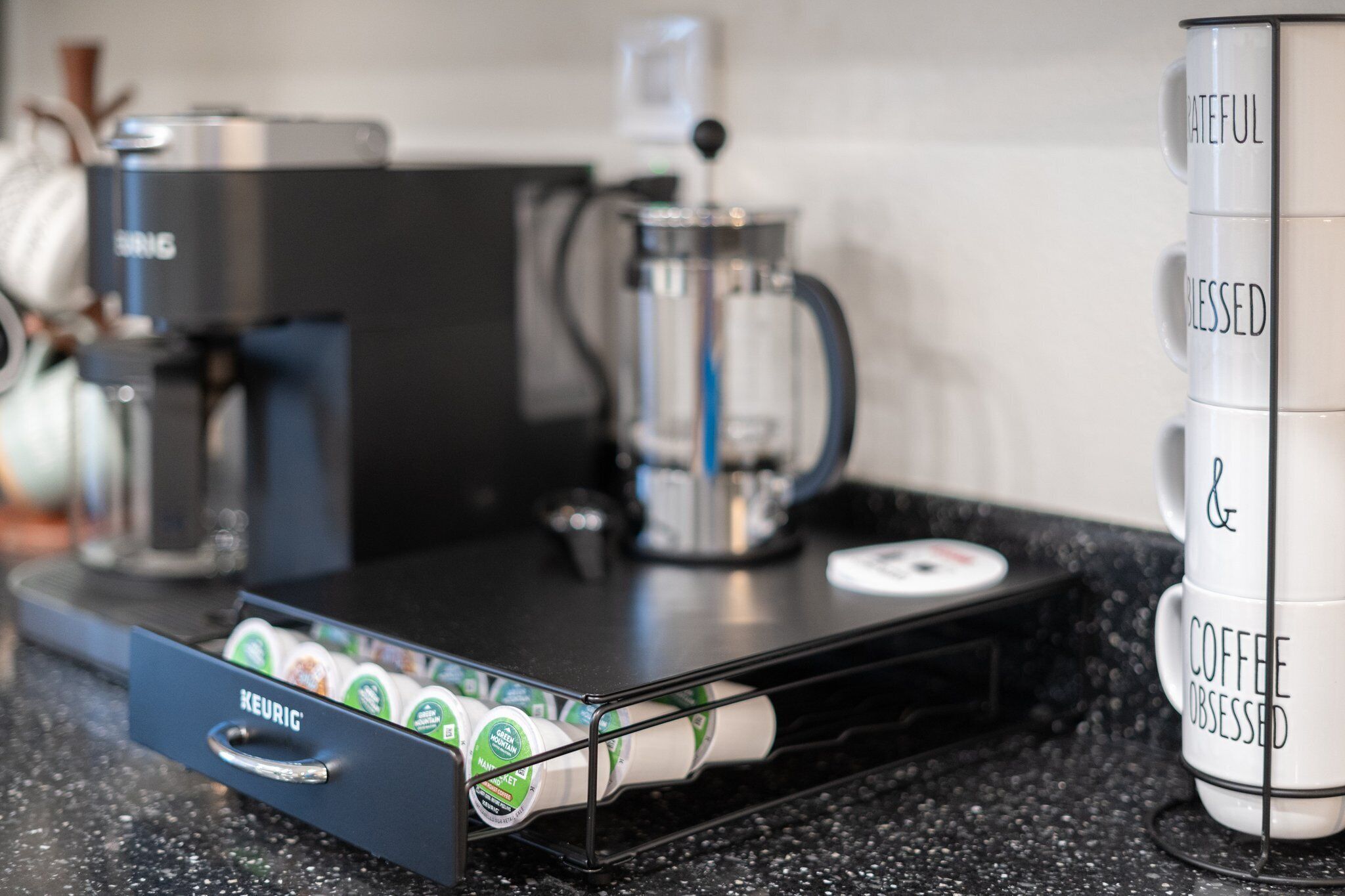 Une machine à café, une machine Keurig et une pile de tasses à café ornées de phrases motivantes.