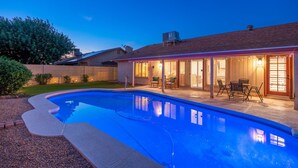 Una piscina con un patio, una zona relax all'aperto coperta e una casa in mattoni sullo sfondo.
