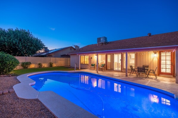 A swimming pool with a patio area, a covered outdoor seating area, and a brick house in the background.