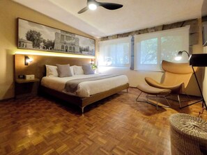 A bedroom with a wooden floor, a bed with pillows, a chair, a lamp, and a painting on the wall.
