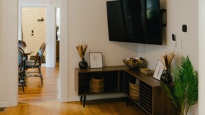 A living room with a flat-screen TV mounted on the wall, a wooden entertainment unit with baskets, and a dining area visible through an open door. - Cozy Updated Century Gem- Prime Location - Sleeps 10- Make yourself at home!  (Cleveland)