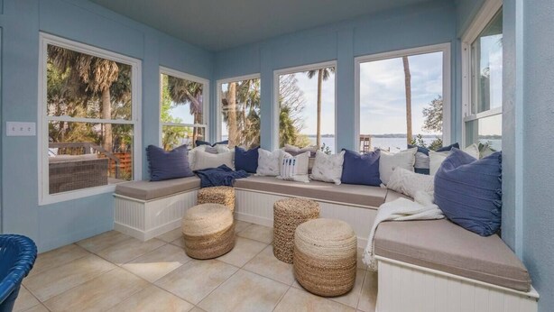 Ein Wintergarten mit Blick auf Palmen und das Meer, ausgestattet mit einer integrierten Bank mit Kissen und gewebten Hocker.