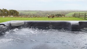 A hot tub with a view of a rural landscape, including stone structures and a wooden fence. - Pol Glas Hall (Helston)