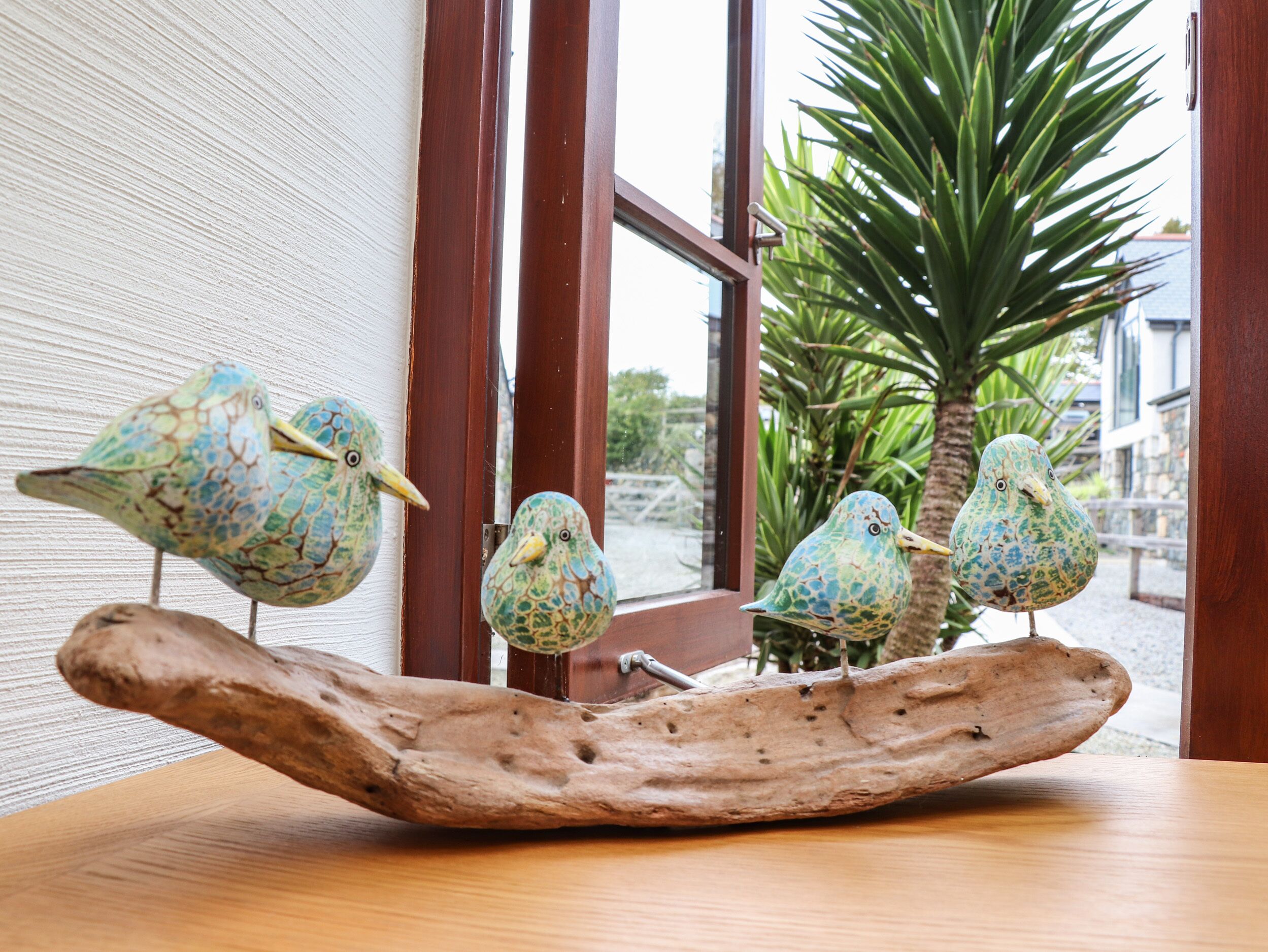 A wooden sculpture with four birds on a branch, placed on a table near a window with a view of a palm tree and buildings.