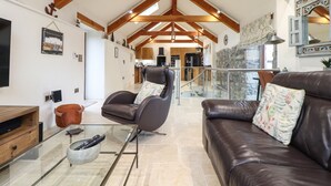 A spacious living area with a high ceiling, wooden beams, a glass coffee table, leather sofas, and a kitchen in the background. - Pol Glas Hall (Helston)