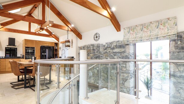 A spacious kitchen with wooden beams, a dining area with a glass railing, and a stone wall with a window. - Pol Glas Hall (Helston)