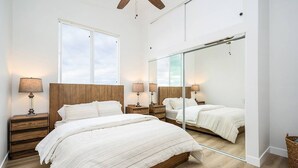 Une chambre avec un grand lit, une tête de lit en bois, des tables de chevet avec des lampes, un grand miroir et une fenêtre donnant sur le ciel.