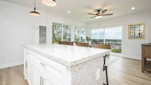 Une cuisine spacieuse dotée d’un grand îlot central, de luminaires suspendus, d’un ventilateur de plafond et de grandes fenêtres offrant une vue sur l’extérieur.