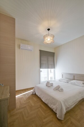 Une chambre d’hôtel avec un grand lit, un plancher en bois, un luminaire au plafond et une fenêtre donnant sur des immeubles.