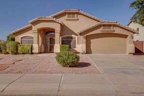 A single-story house with a beige facade, a front garage, and a small front yard with a gravel driveway and a trimmed bush.