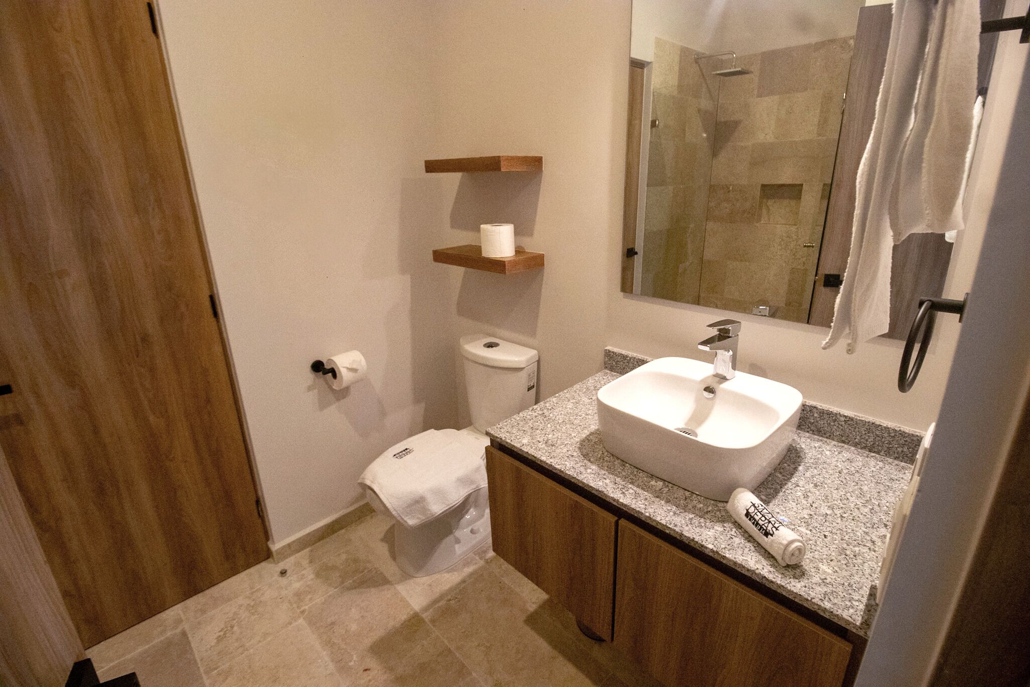 A bathroom with a sink, toilet, and shower. There are wooden shelves above the toilet and a mirror above the sink.