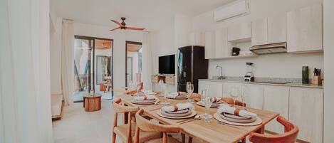 A dining area with a wooden table set for four, a ceiling fan, a black refrigerator, and a kitchen counter with a blender.
