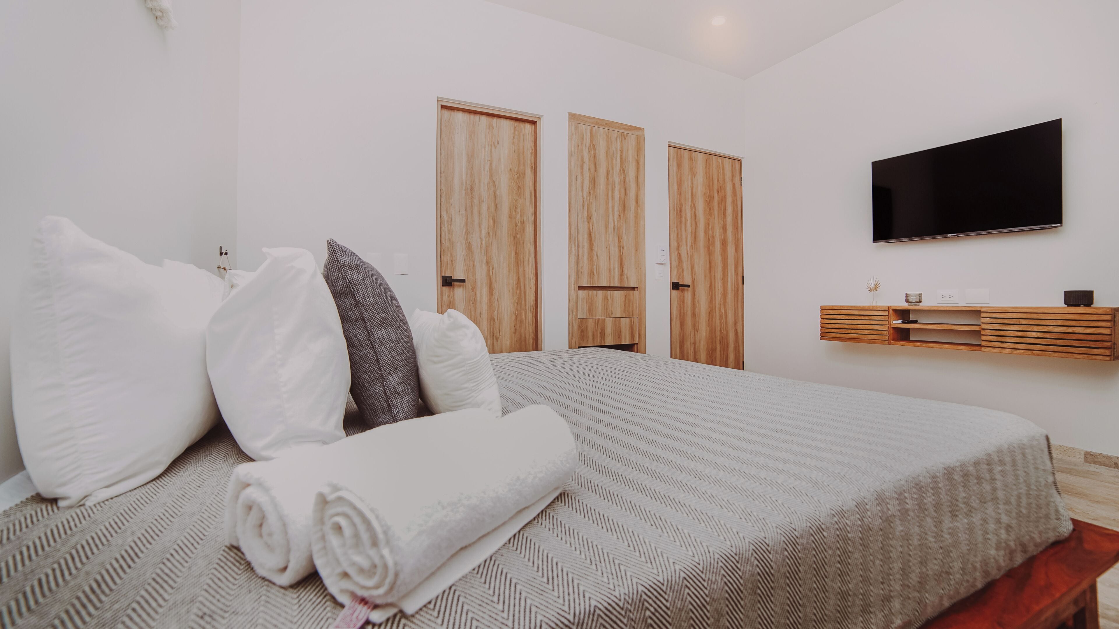 Una camera da letto con un letto, cuscini, televisione, un'armadio in legno e una mensola.