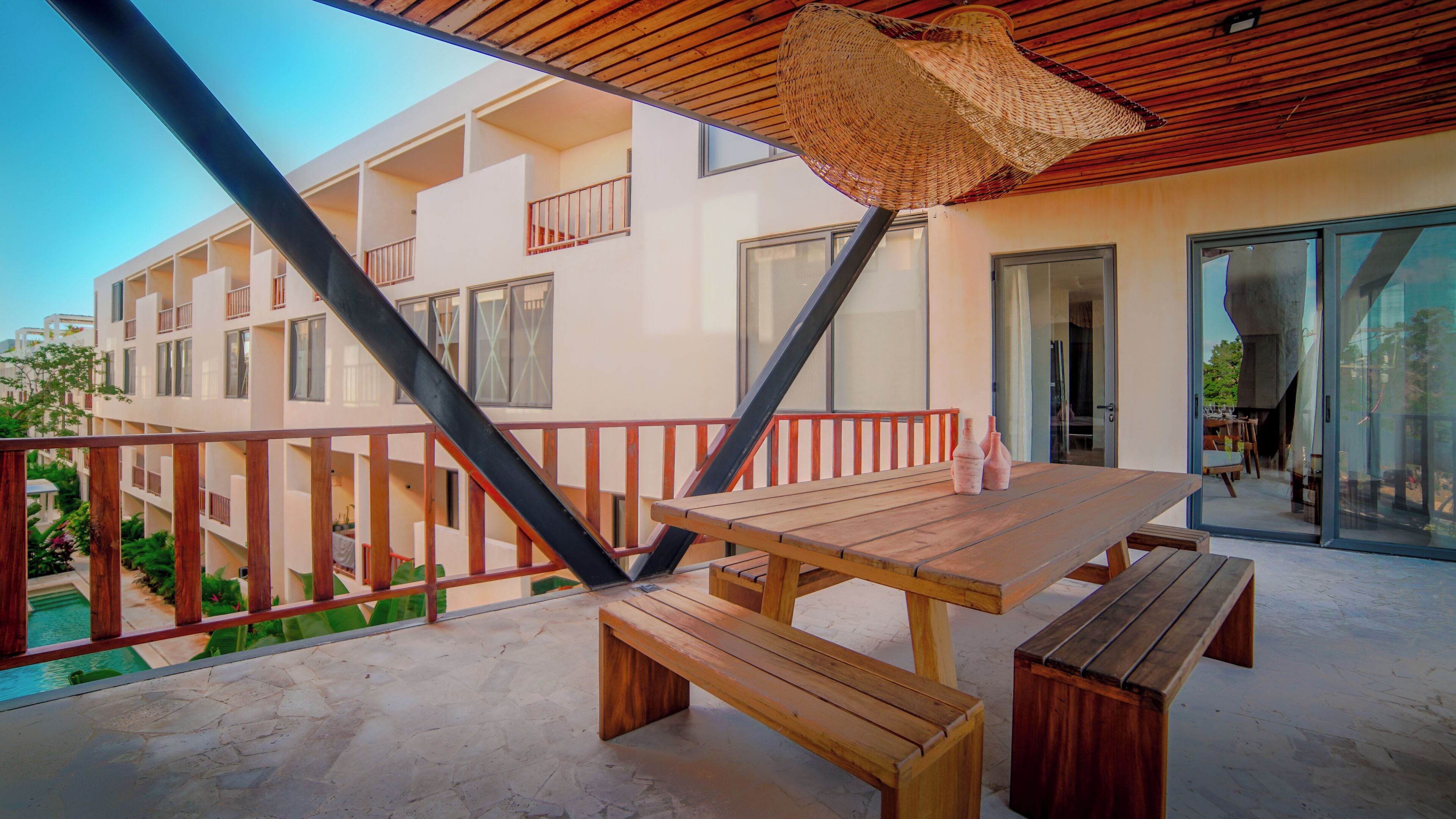 Ein Balkon mit Holztisch und Bänken, einer gewebten Decke sowie einem Gebäude mit mehreren Balkonen und großen Fenstern.