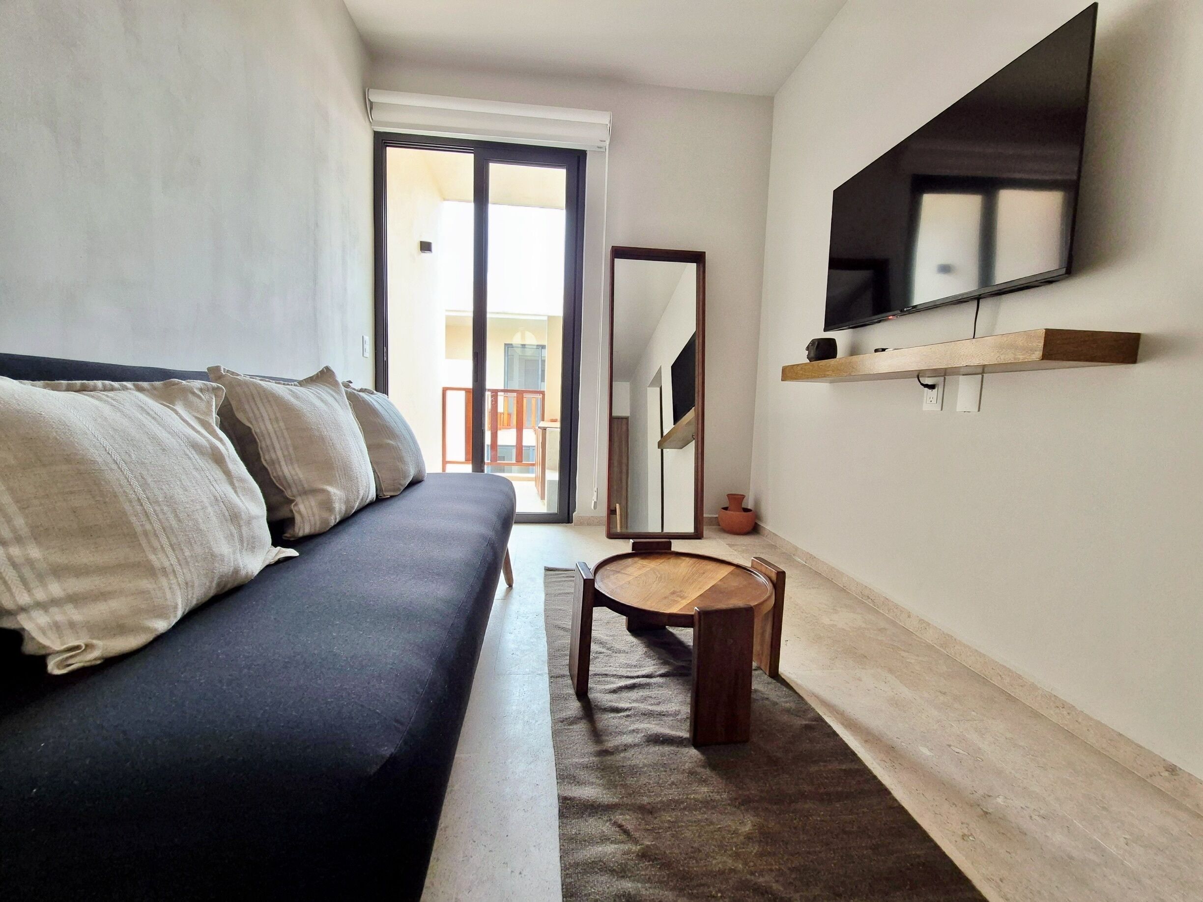 A modern living room with a sofa, a round wooden table, a mirror, and a TV mounted on the wall.