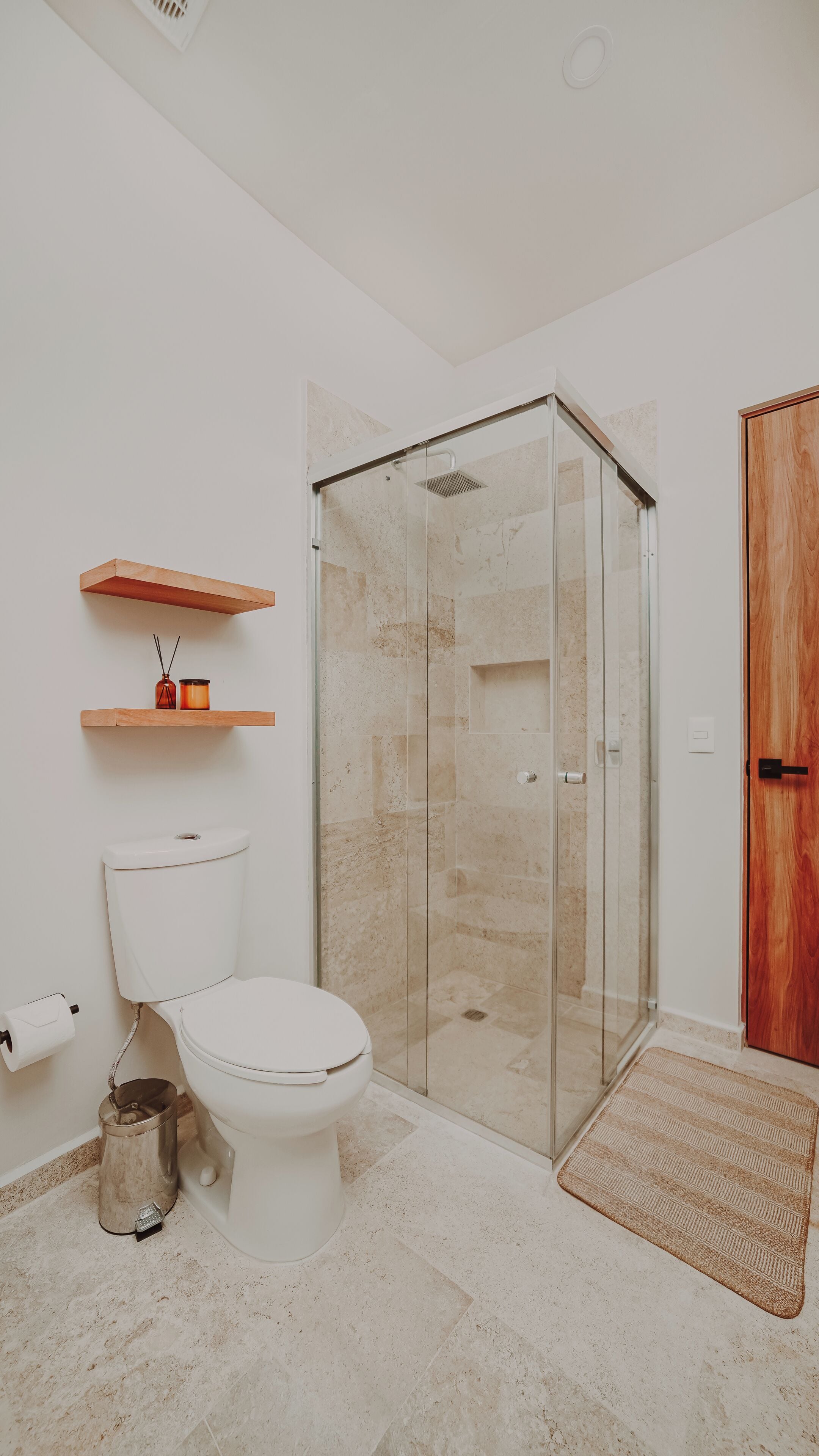 Een badkamer met een toilet, een glazen douchecabine, een houten deur en een klein plankje met een kaars en een vaas.