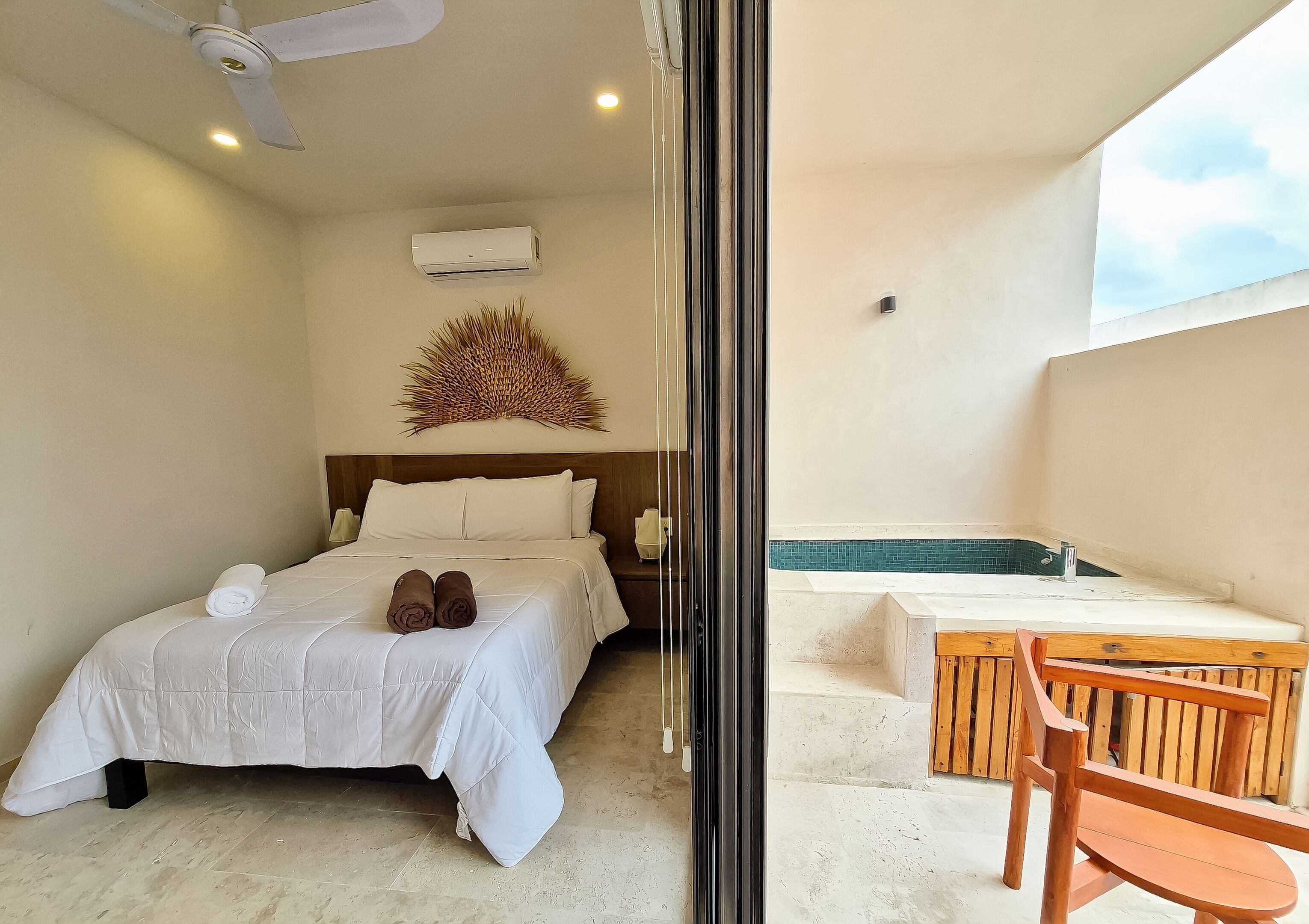 Camera d'hotel con un letto, una ventola a soffitto, un condizionatore d'aria, un elemento decorativo al muro e vista su un'area benessere con una sedia di legno e una vasca da bagno.