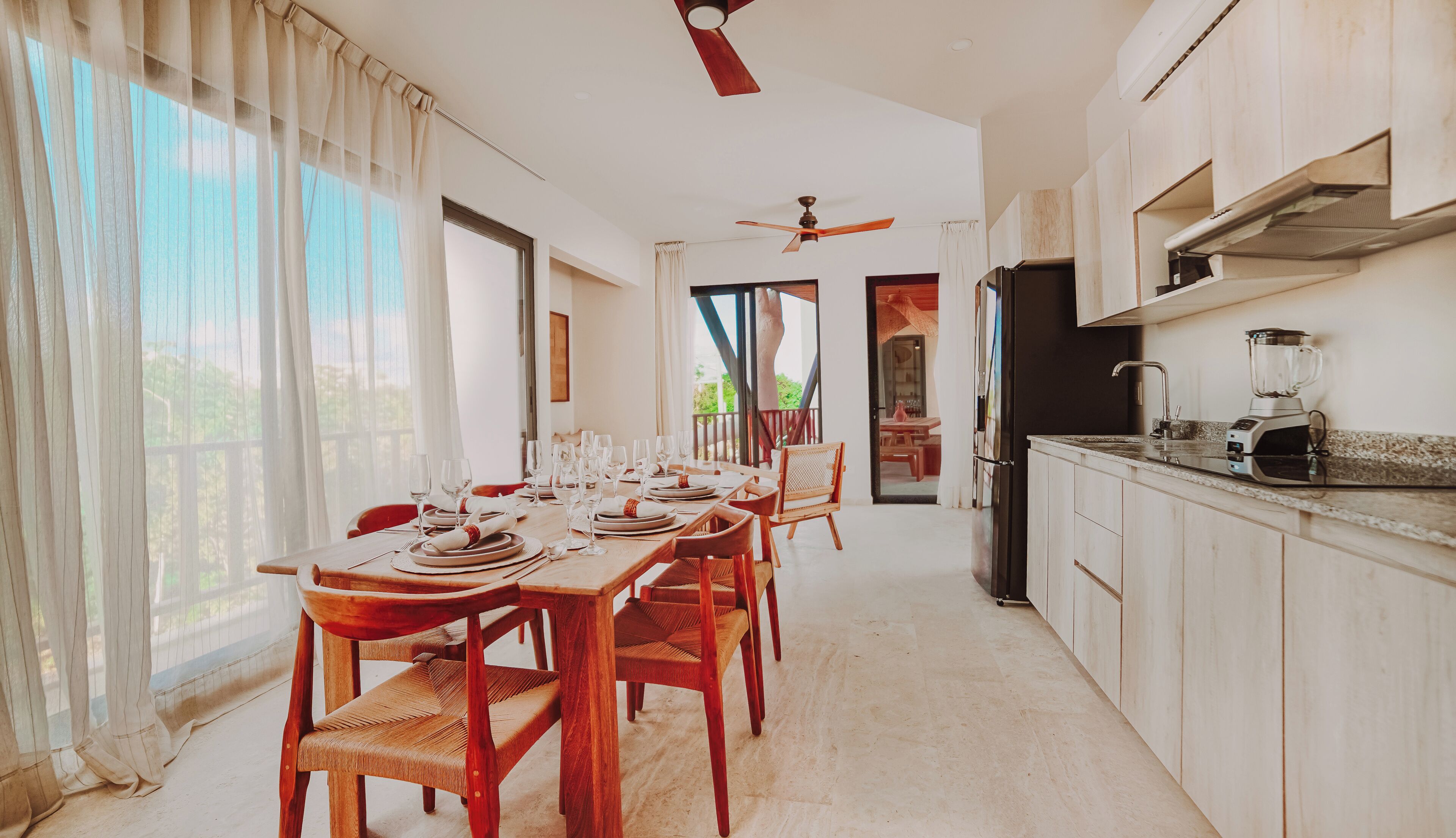 A dining area with a table set for six, wooden chairs, a ceiling fan, and a kitchen with a refrigerator and blender.