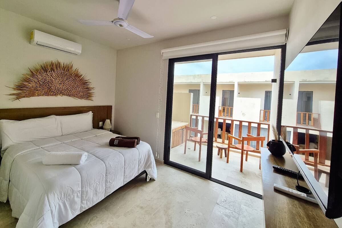 A hotel room with a bed, a ceiling fan, a large window, and a balcony with chairs.