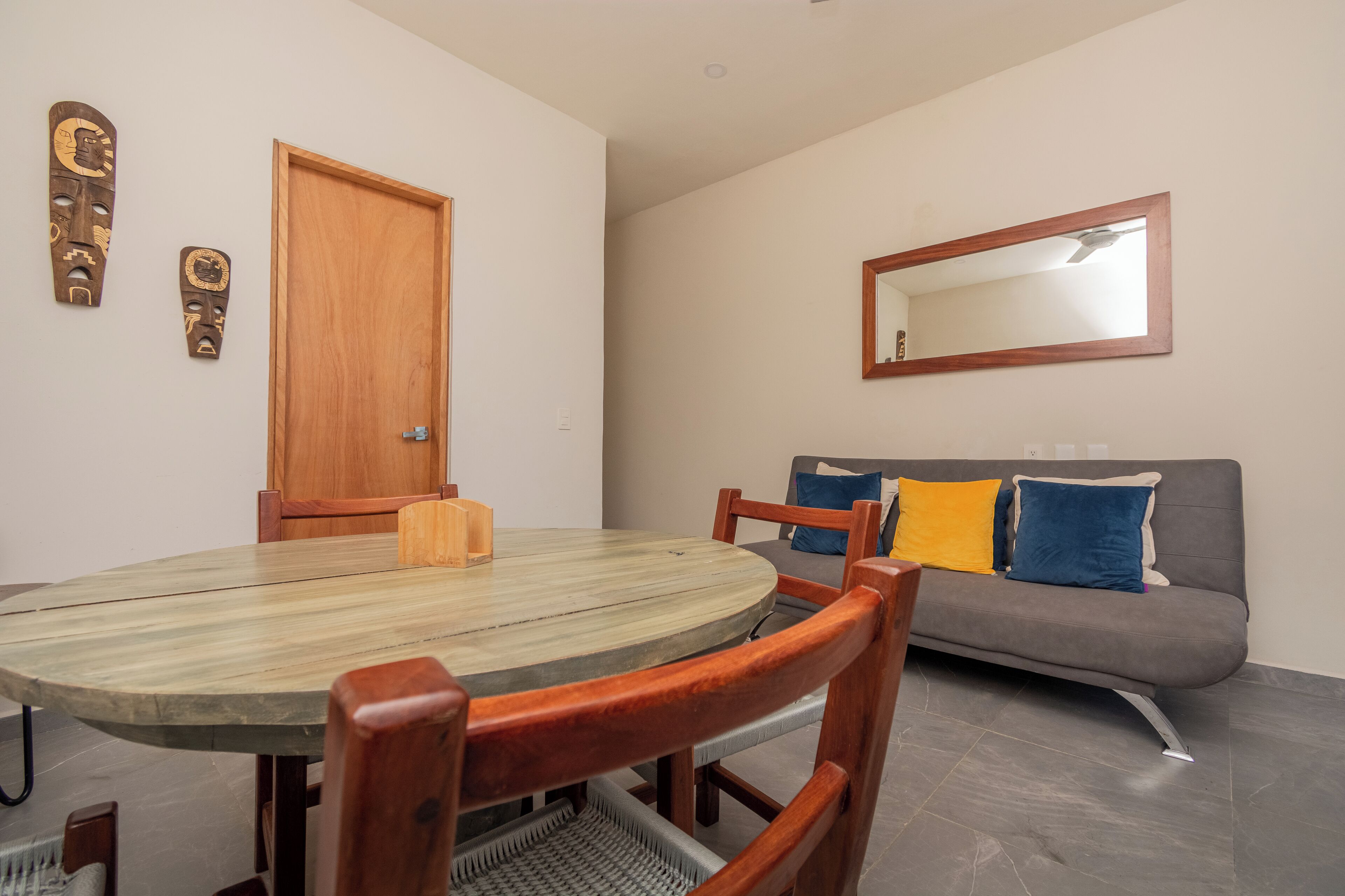 A dining area with a round table and chairs, a wooden door, a gray sofa with cushions, and a mirror on the wall.