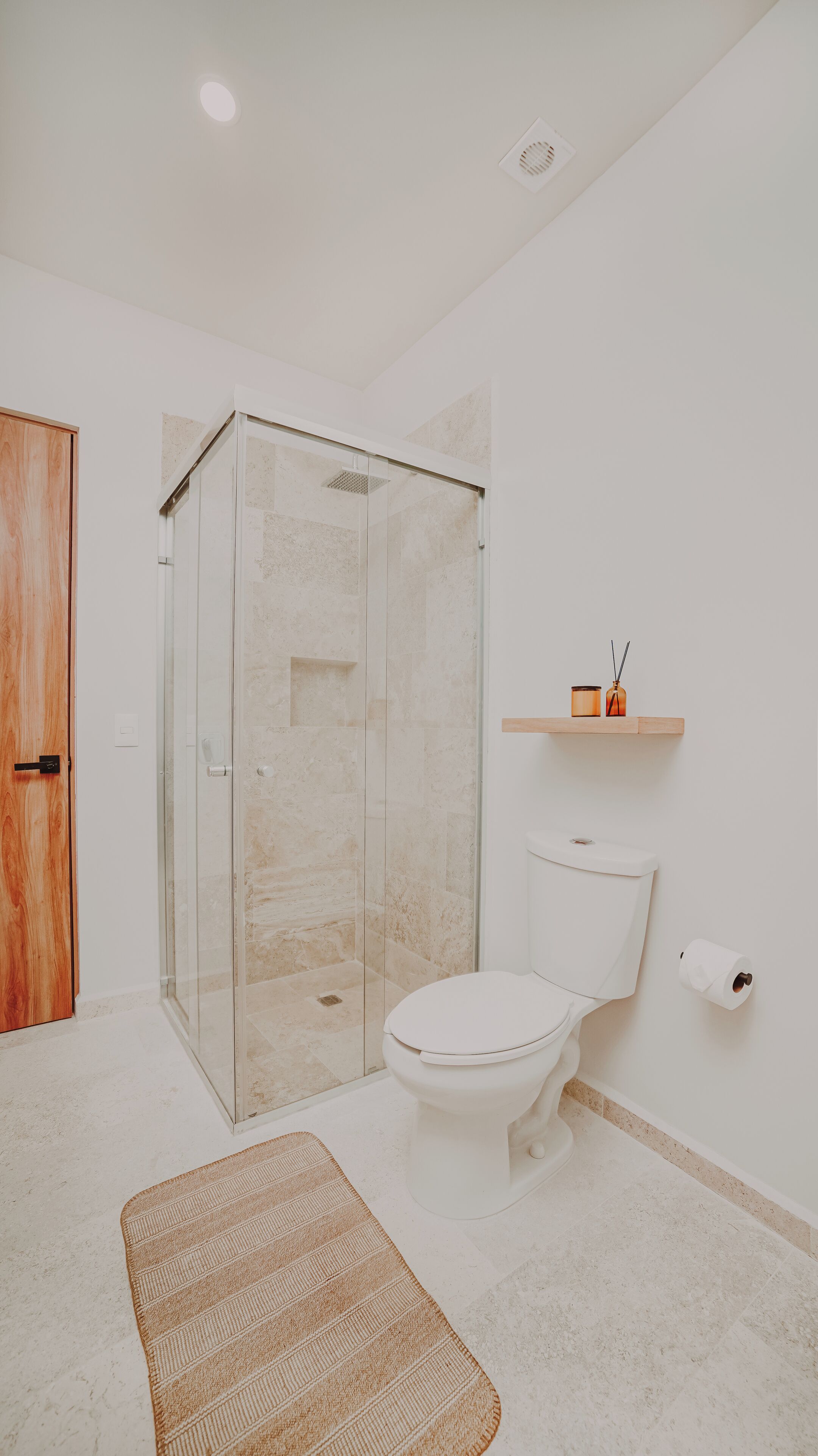 A bathroom with a glass-enclosed shower, a toilet, a wooden door, and a small shelf with a candle.