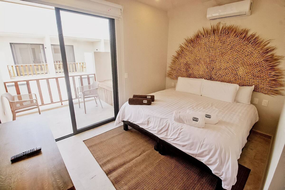A bedroom with a bed, a small table, a chair, a large window, and a balcony.