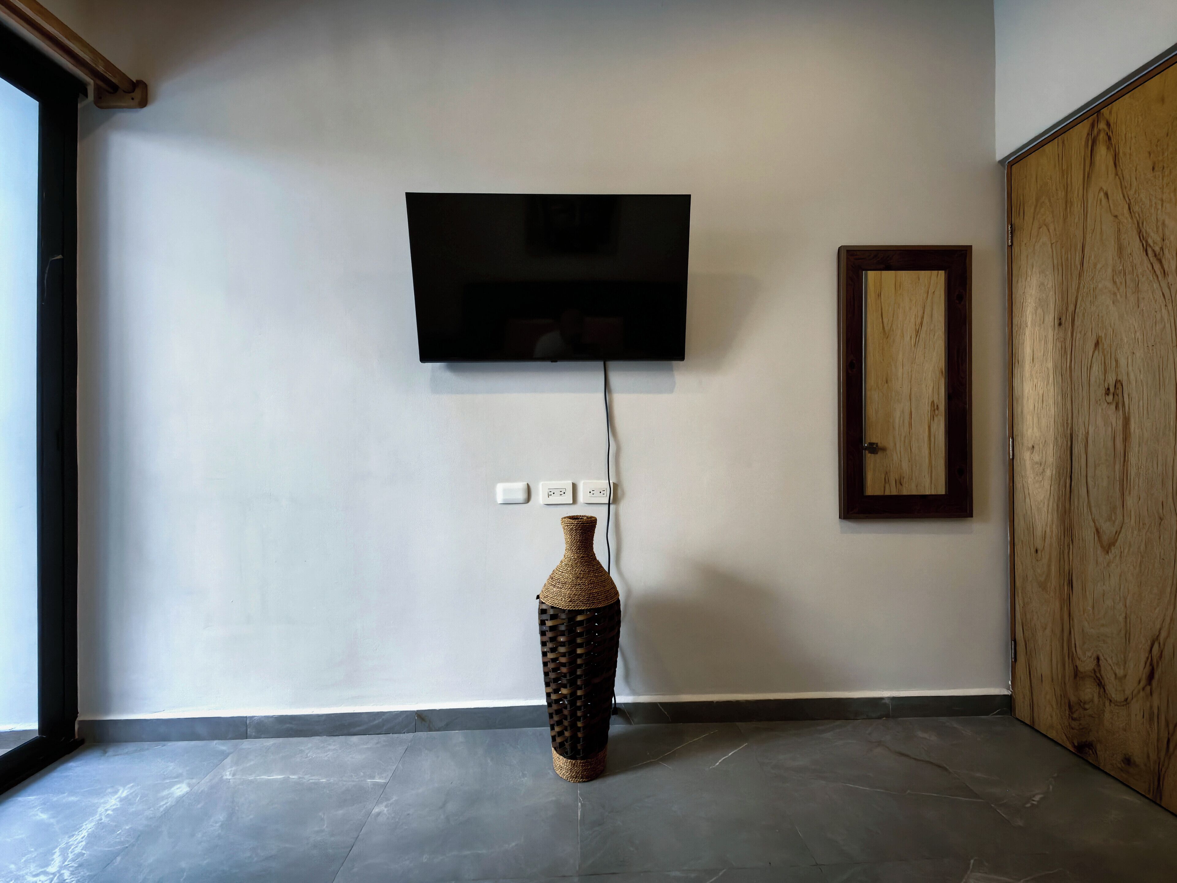 A room with a flat-screen TV mounted on the wall, a wooden door, and a woven vase on the floor.