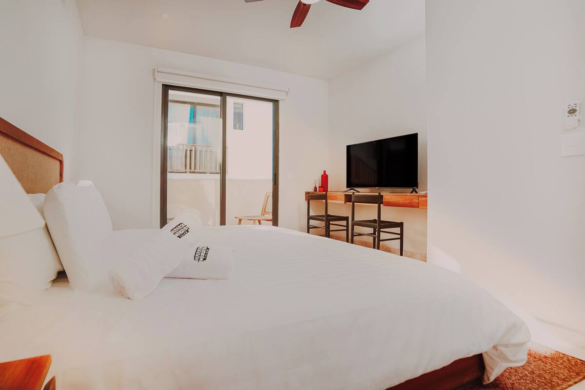 A hotel room with a bed, a TV, a balcony with a chair, and a wooden desk.